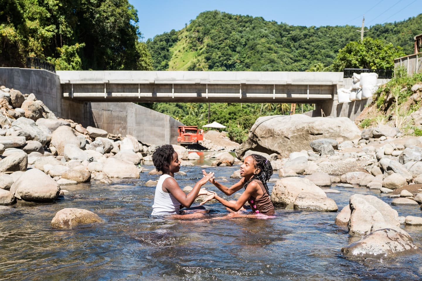 Two girls playing in a shallow river in front of a new concrete road bridge, with forested hills behind